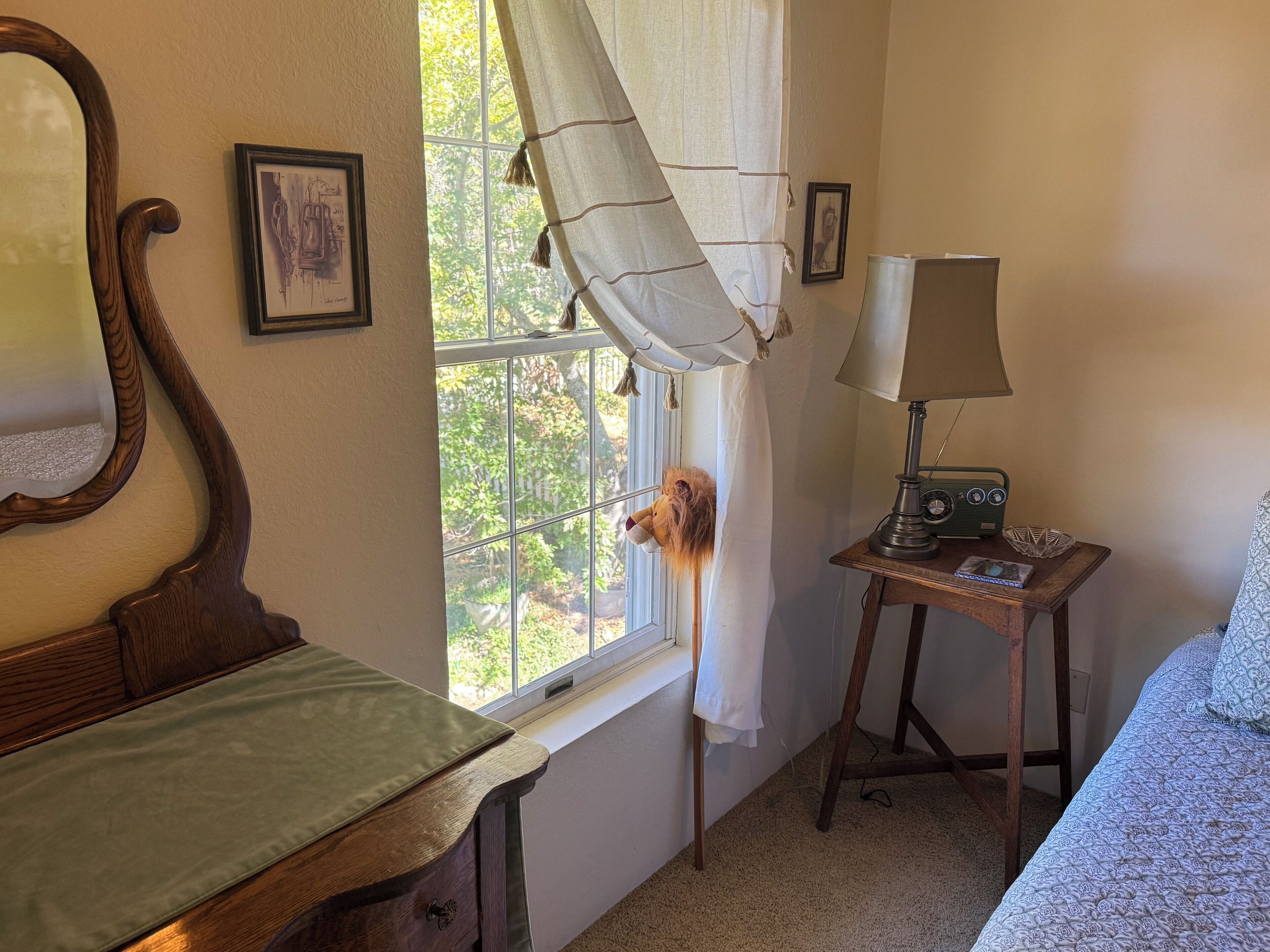 A room with a wooden bed, a small table with a lamp and radio, a window with curtains, and a framed picture on the wall.