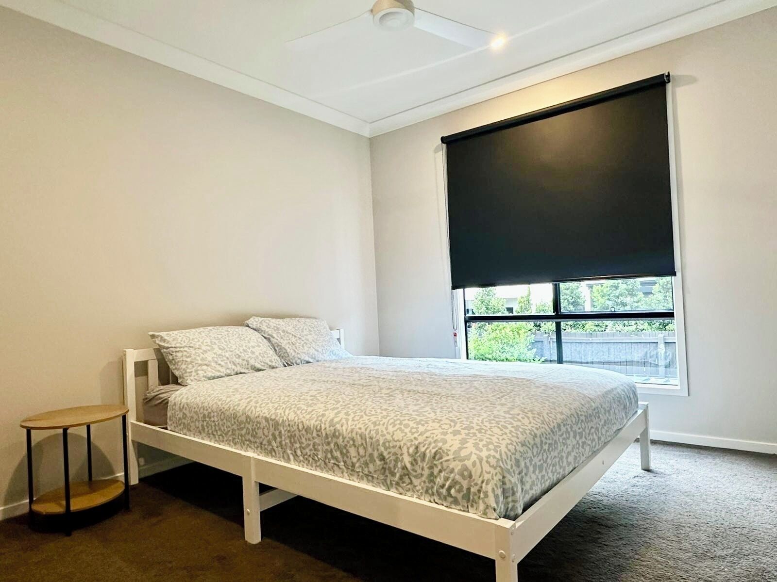 Une chambre à coucher avec un lit, une petite table, une fenêtre avec un store noir et un ventilateur de plafond.
