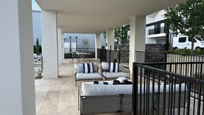 A covered outdoor seating area with two lounge chairs and a table, surrounded by a black metal railing.