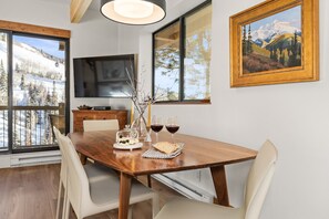 A dining area with a wooden table, chairs, a TV, and a painting on the wall. - Club-B211: Fully Renovated, Panoramic Mountain Views, Prv W/D, Ski In Ski Out (Steamboat Springs)