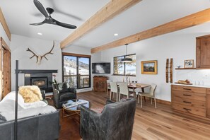 A spacious living room with a fireplace, a large window with a ski view, a dining area, and wooden beams on the ceiling. - Club-B211: Fully Renovated, Panoramic Mountain Views, Prv W/D, Ski In Ski Out (Steamboat Springs)