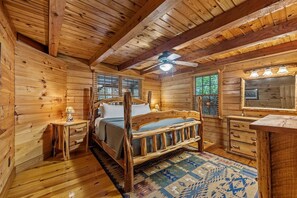 A wooden cabin bedroom with a bed, a nightstand, a dresser, a ceiling fan, and a window with blinds. - Toccozy Cabin Retreat - Riverfront | Pet Friendly | Hot Tub | Game Room | Firepit (Mineral Bluff)