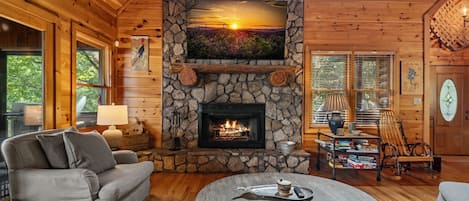 A cozy cabin interior with a stone fireplace, wooden furniture, and a large window.