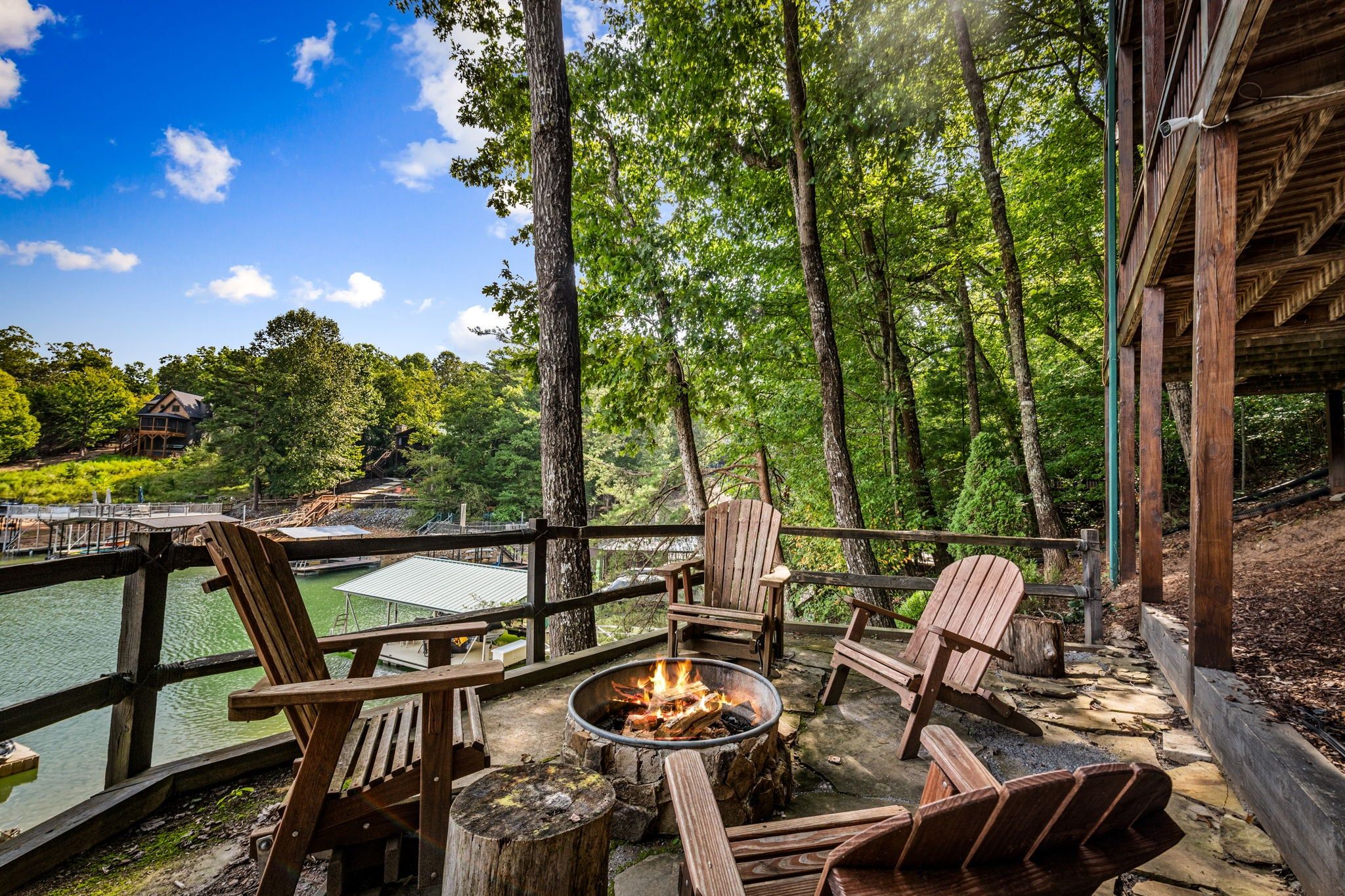 A wooden deck with Adirondack chairs, a fire pit, and a view of a lake and forest.