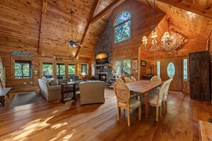 Une cabane en bois spacieuse, dotée d’un plafond haut, d’une cheminée, d’un coin repas et de grandes fenêtres.