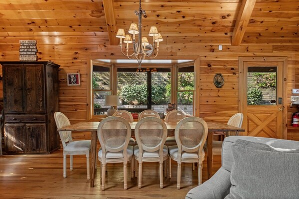 A wooden cabin interior with a dining table surrounded by wicker chairs, a chandelier, and a cozy seating area with a sofa. - Johnny's Lakeside Hideaway - Private Dock | Hot Tub | Game Room (Morganton)