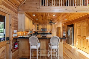 A wooden cabin kitchen with a central island, two bar stools, a ceiling fan, and a stainless steel refrigerator. - Johnny's Lakeside Hideaway - Private Dock | Hot Tub | Game Room (Morganton)