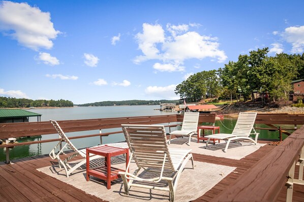Terraza de madera con sillones de descanso y una pequeña mesa, con vista a un lago tranquilo y un cielo despejado.