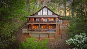 A two-story log cabin with a balcony and multiple windows, surrounded by dense forest. - It's All About the Views - Mtn Views | Hot Tub | Pool Table | Arcade Games | Fire Pit (Blue Ridge)