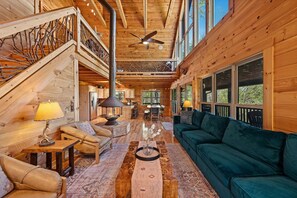 A log cabin interior with a wooden staircase, a ceiling fan, a dining area, and a green sofa. - It's All About the Views - Mtn Views | Hot Tub | Pool Table | Arcade Games | Fire Pit (Blue Ridge)