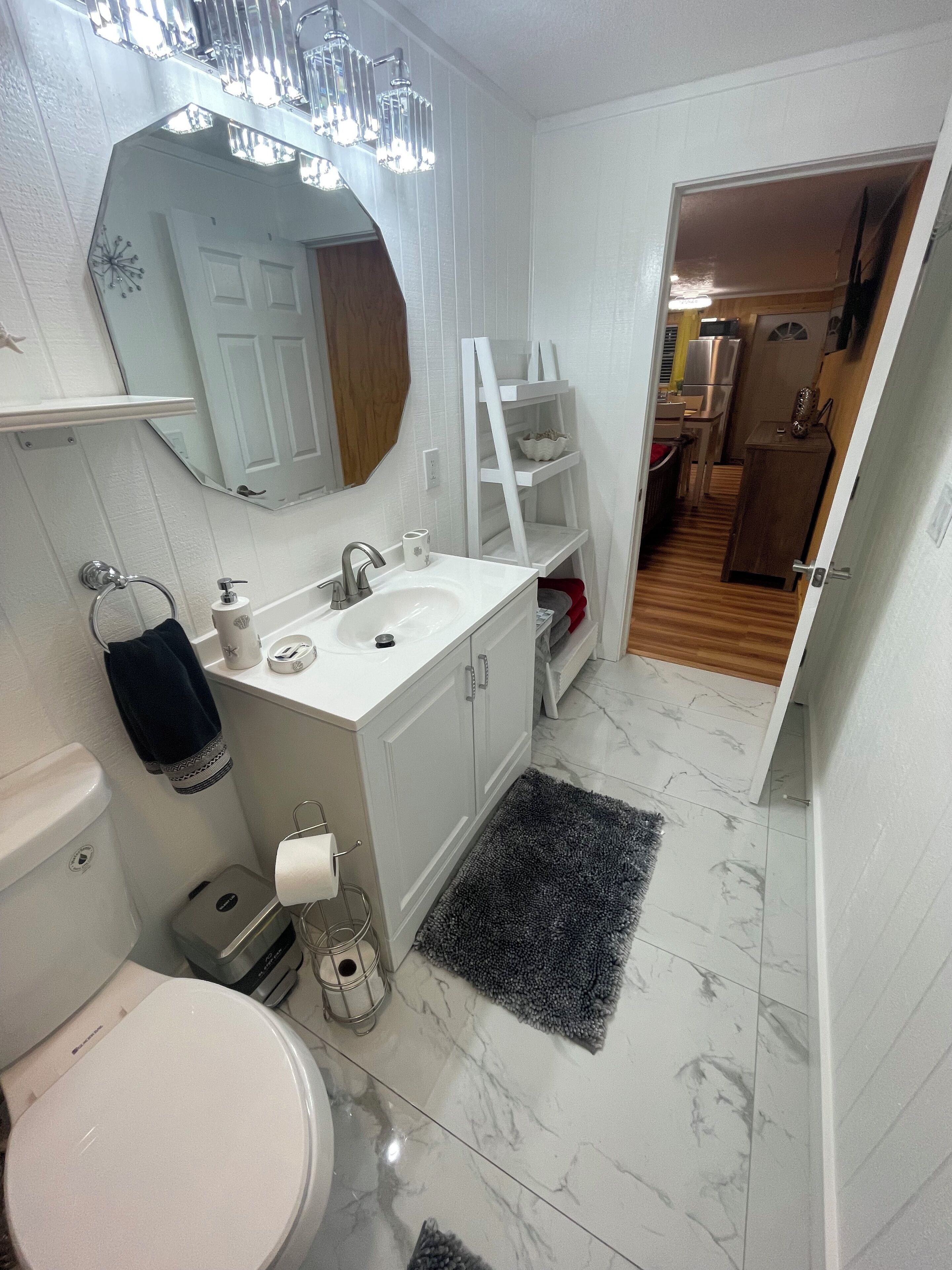 Un baño moderno con un lavamanos, espejo y retrete blancos. Hay una alfombra negra en el suelo y un estante blanco con artículos de aseo.