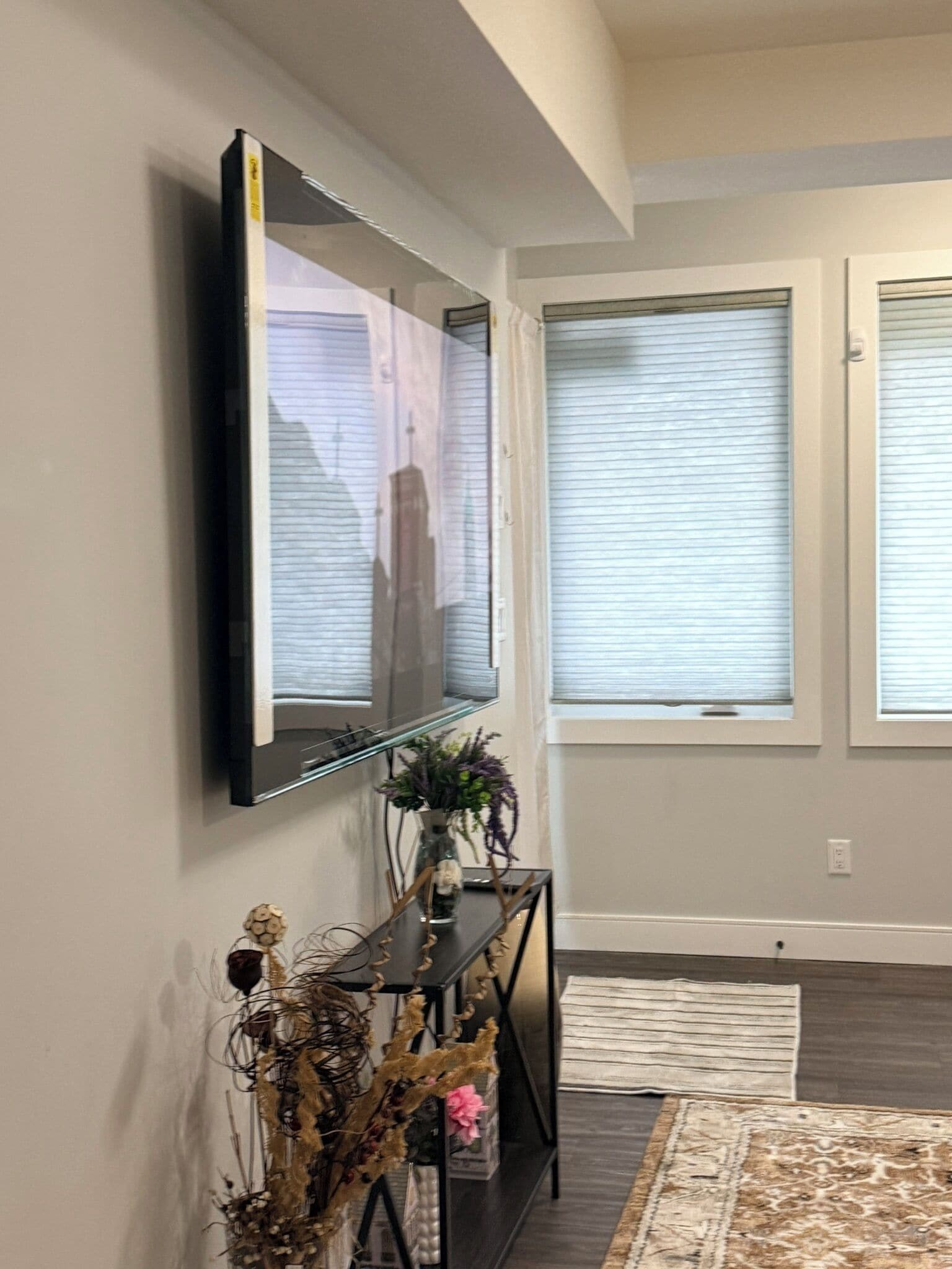 Un salon doté d’un téléviseur à écran plat fixé au mur, d’une console sur laquelle sont disposés des objets décoratifs, d’un tapis à motifs et de grandes fenêtres équipées de stores.