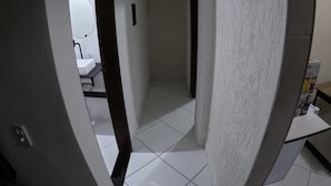 A hallway with a white tiled floor, a door leading to a bathroom with a white sink and mirror, and a small table with a box on it. - Practical and cozy house in Vila Velha (2 guests) (Vila Velha)