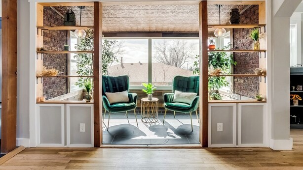 Quarto com duas poltronas verdes, uma mesa redonda pequena e prateleiras com plantas e objetos decorativos.