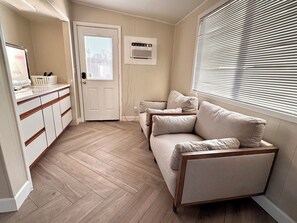 Ein Zimmer mit Fischgrätparkett, einem beigen Sofa, einer weißen Tür und einem Fenster mit Jalousien.