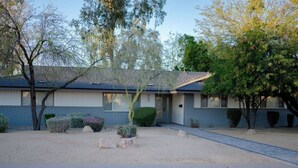 A two-story building with a front entrance, a driveway, and a small garden area with various shrubs and a tree. - Golf House Retreat-Newly Remodeled 4BR (Tempe)