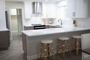 A modern kitchen with white cabinetry, a stainless steel range hood, a built-in oven, a large island with bar stools, and a sink. - Golf House Retreat-Newly Remodeled 4BR (Tempe)
