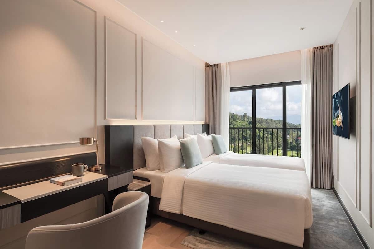 A hotel room with a bed, a desk, a chair, a TV, and a view of greenery.