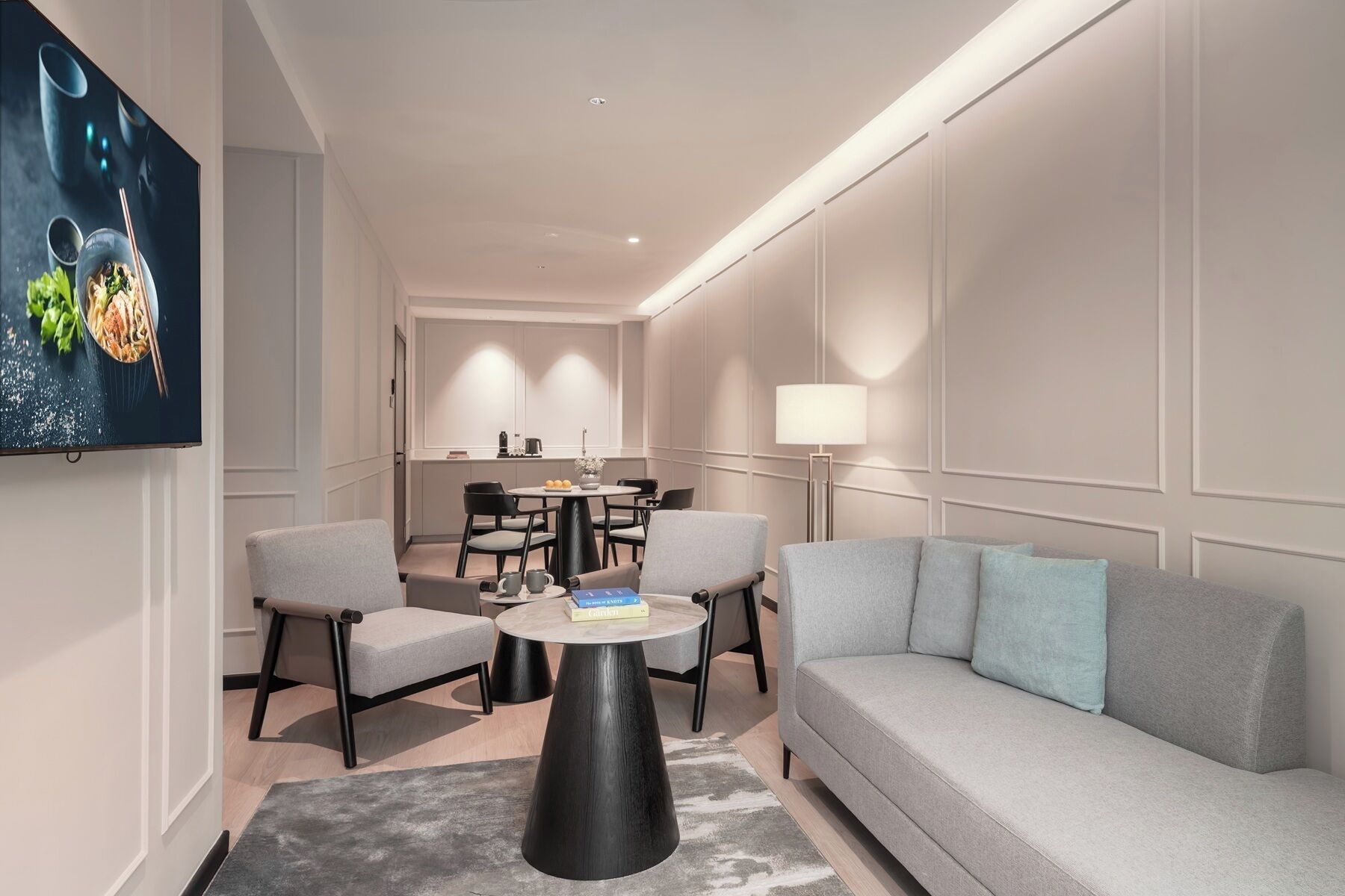 Une chambre d’hôtel moderne avec un canapé gris, deux fauteuils, une table basse ronde, un coin repas avec une table et des chaises, un téléviseur fixé au mur et une lampe sur pied.