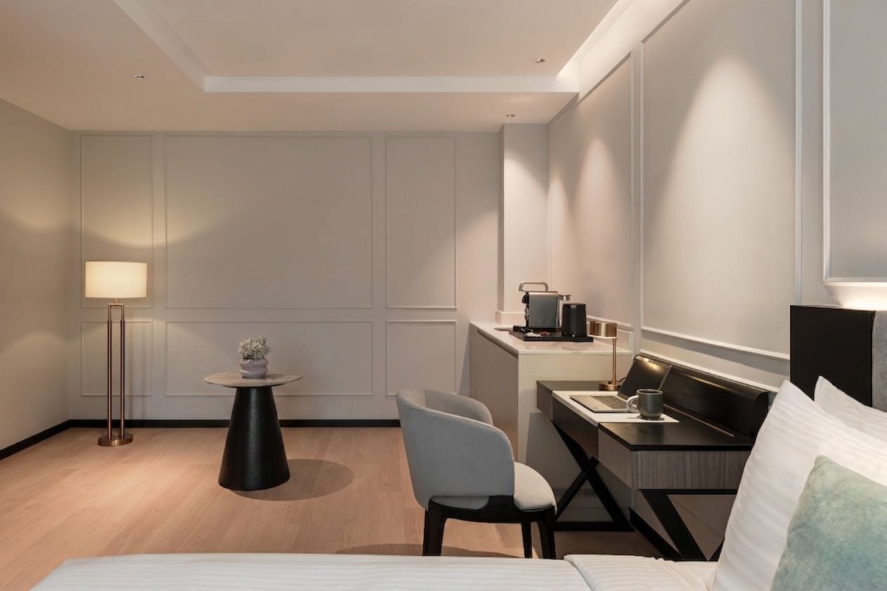 A modern hotel room with a bed, a desk with a laptop, a chair, a small round table with a lamp, and a potted plant.