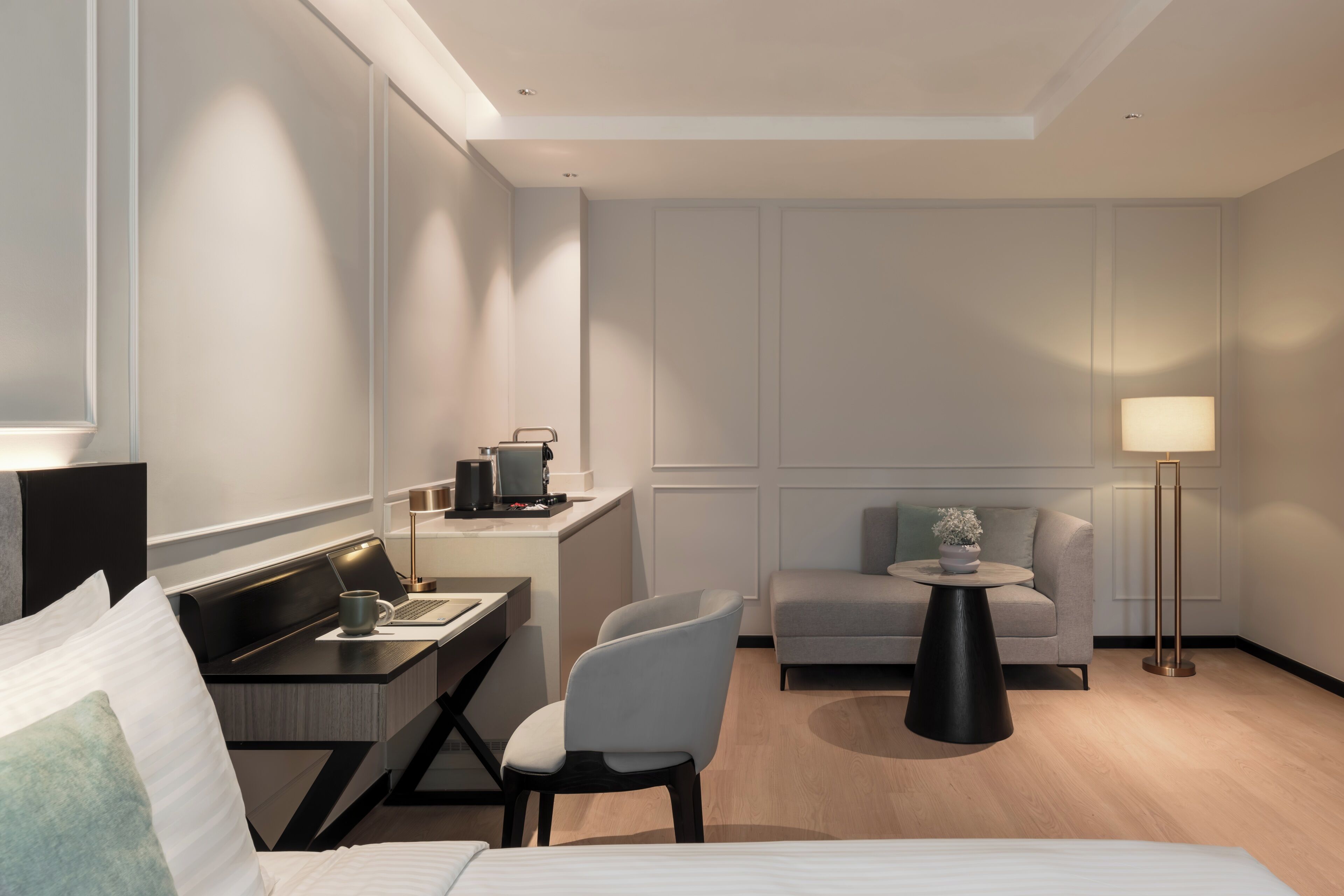A modern hotel room with a bed, desk, chair, sofa, and a small table with a lamp.