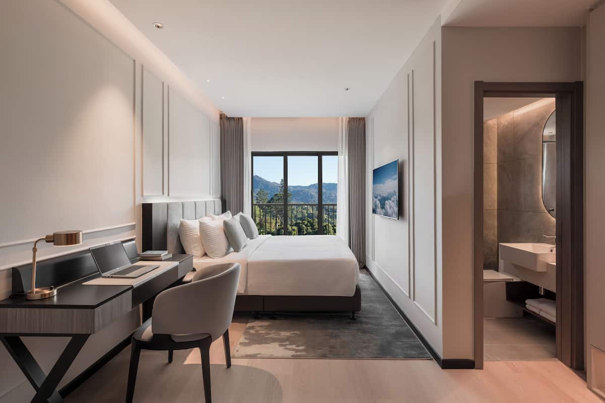 A modern hotel room with a bed, desk, chair, and a view of the outdoors.