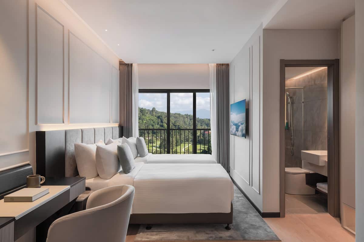 A modern hotel room with a large bed, a desk, a chair, a bathroom, and a balcony with a view of the greenery.