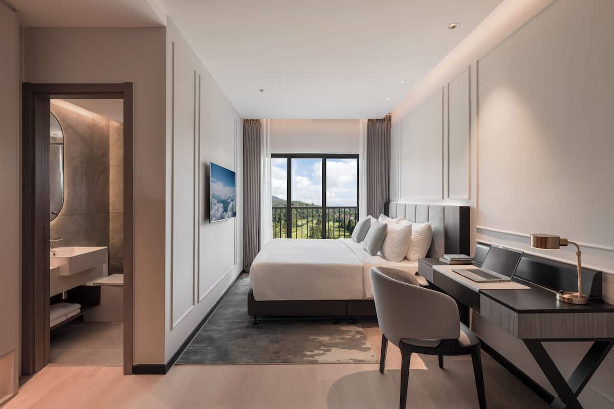 A modern hotel room with a bed, desk, chair, and a view of the outdoors.