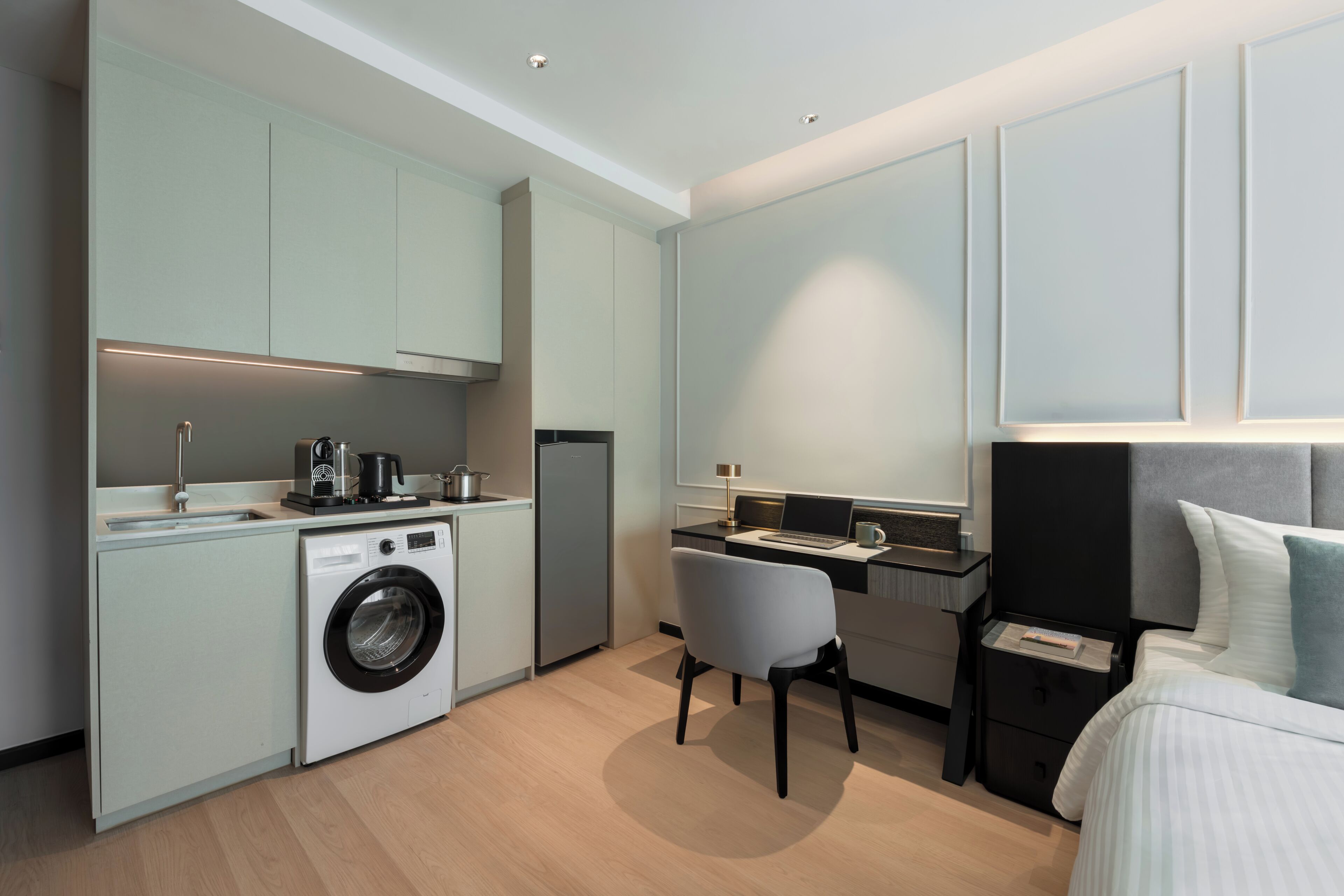 A compact hotel room with a kitchenette, a washing machine, a desk with a chair, and a bed with a headboard.