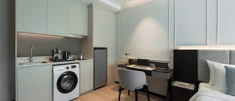 A compact hotel room with a kitchenette, a washing machine, a desk with a chair, and a bed with a headboard.