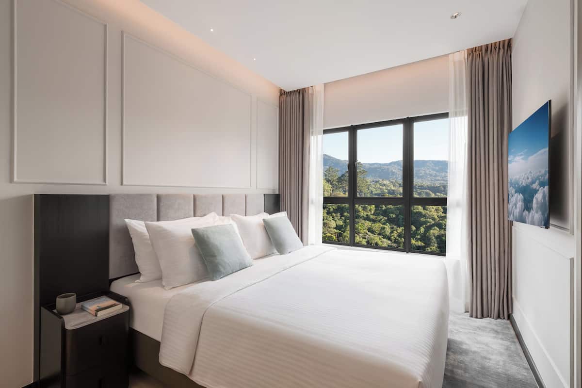 A bedroom with a large bed, a nightstand, a TV, and a window with a view of greenery.