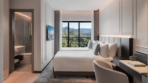 A modern hotel room with a large bed, a desk, a chair, a bathroom, and a balcony with a view of the mountains.