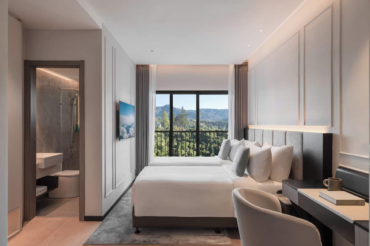 A modern hotel room with a large bed, a desk, a chair, a bathroom, and a balcony with a view of the mountains.