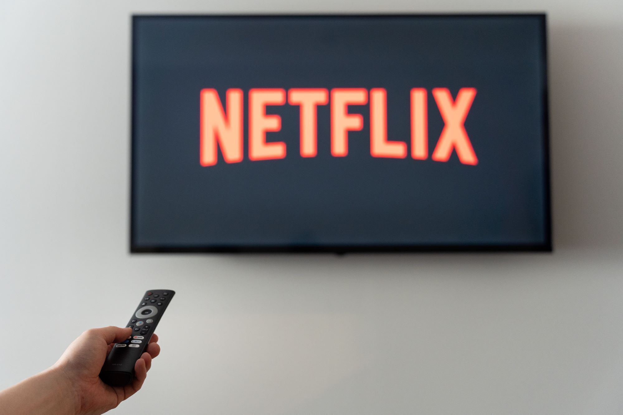Käsi pitää kaukosäädintä television edessä, ja televisiossa näkyy punaisilla kirjaimilla sana »NETFLIX».