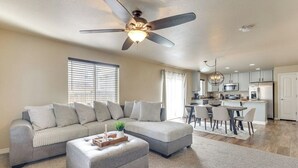 A spacious living room with a grey sectional sofa, ottoman, and a ceiling fan. A dining area with a table and chairs is visible in the background.