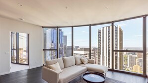 Un salon moderne avec un canapé d’angle beige, une petite table ronde et une vue sur le paysage urbain grâce à de grandes fenêtres.