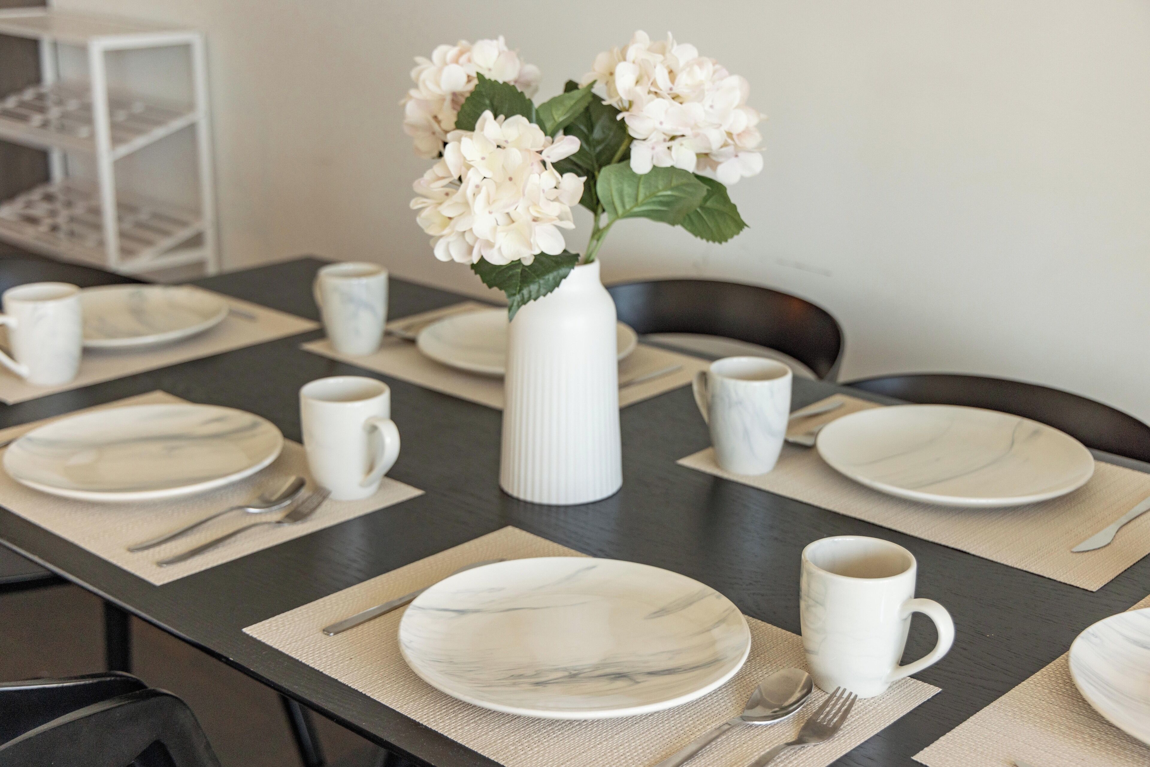 Una mesa de comedor dispuesta con platos blancos, cubiertos y tazas blancas, con un jarrón blanco con flores blancas en el centro.