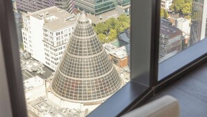 Un immeuble de grande hauteur doté d’une structure conique en verre remarquable, vu à travers une fenêtre avec un canapé blanc et un cadre noir.