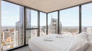 Une chambre d’hôtel moderne avec un grand lit, du linge de lit blanc et une vue sur la ville grâce à de grandes fenêtres du sol au plafond.
