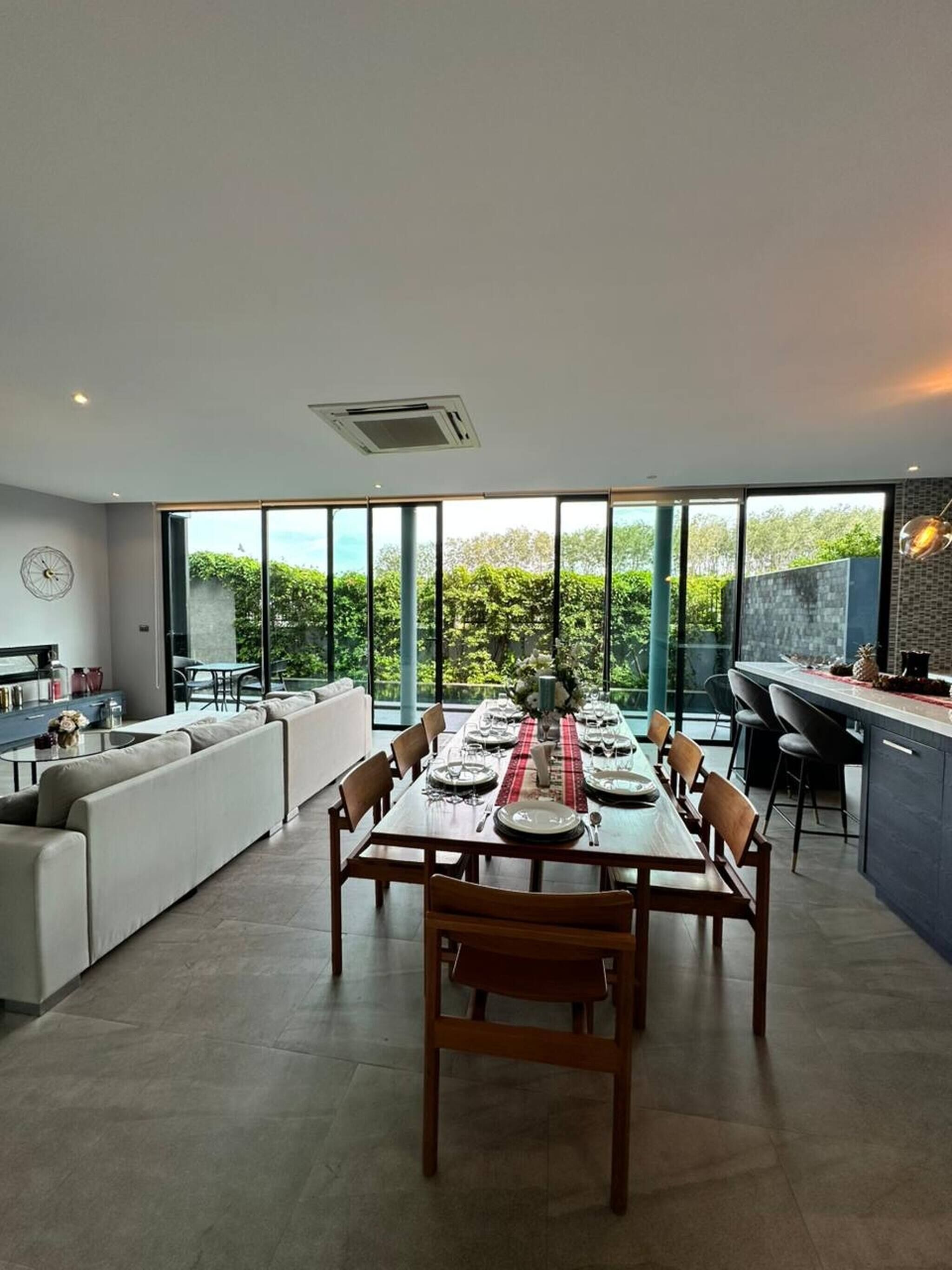 A modern dining area with a large table set for six, surrounded by chairs. There is a sofa and a kitchen area with a bar to the right.