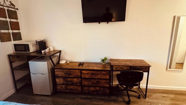 Una habitación compacta con un escritorio de madera, un refrigerador pequeño, un microondas, una silla de oficina negra y un televisor montado en la pared.