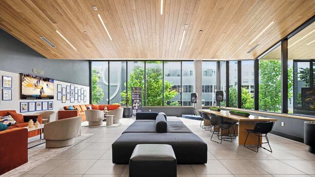 Un área de descanso moderna con techo de madera, ventanas amplias y un conjunto de sofás junto a una barra.