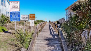 Une promenade en bois mène à une plage où des panneaux indiquent qu’il est interdit de consommer de l’alcool, que les chiens doivent être tenus en laisse et que les chemins sont accessibles aux personnes à mobilité réduite.