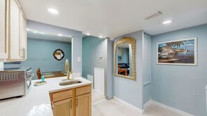 Une cuisine avec un plan de travail blanc, des armoires en bois, une hotte de cuisine en acier inoxydable et un tableau représentant un paysage côtier accroché au mur.