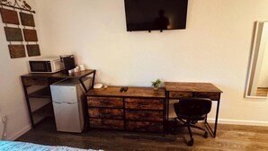 A compact room with a wooden desk, a small refrigerator, a microwave, a black office chair, and a TV mounted on the wall. - 16 - Scenic route stays (Challenge-Brownsville)