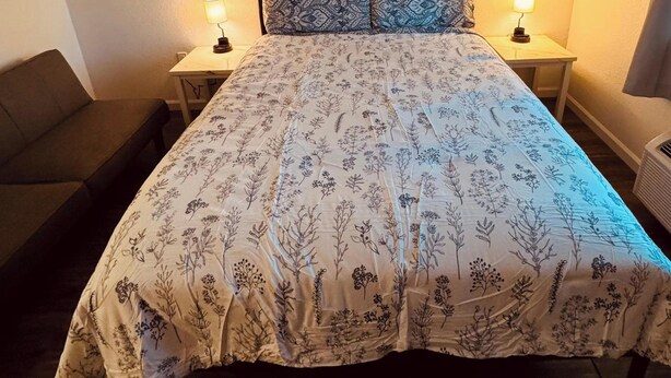 A bed with a floral-patterned bedspread, two bedside tables with lamps, a couch, and a window with a curtain. - 16 - Scenic route stays (Challenge-Brownsville)