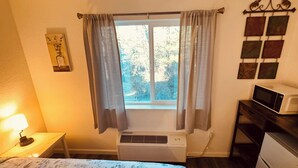 A room with a bed, a bedside lamp, a window with curtains, an air conditioner, a microwave, and a refrigerator. - 16 - Scenic route stays (Challenge-Brownsville)