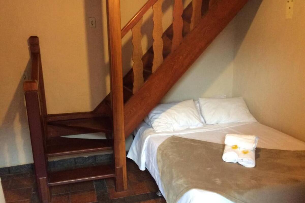 Una cama individual con ropa blanca y una manta marrón, una silla de madera y una escalera de madera.