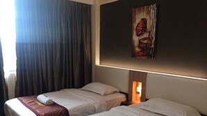A hotel room with two beds, each with a white bedspread and a brown headboard. There is a painting above the beds and a window with curtains. - Hotel Casamas (Malacca City)