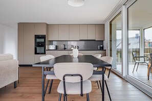 A modern kitchen with a dining area, featuring a large table and chairs, a built-in oven, and a view through large windows. - Blueground | Oerlikon, balcony, nr Zurich Park (Zurich)
